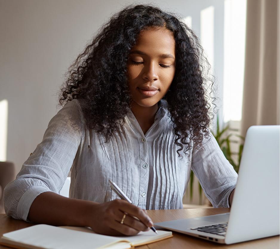 woman writing in a notebook while working on a laptop.